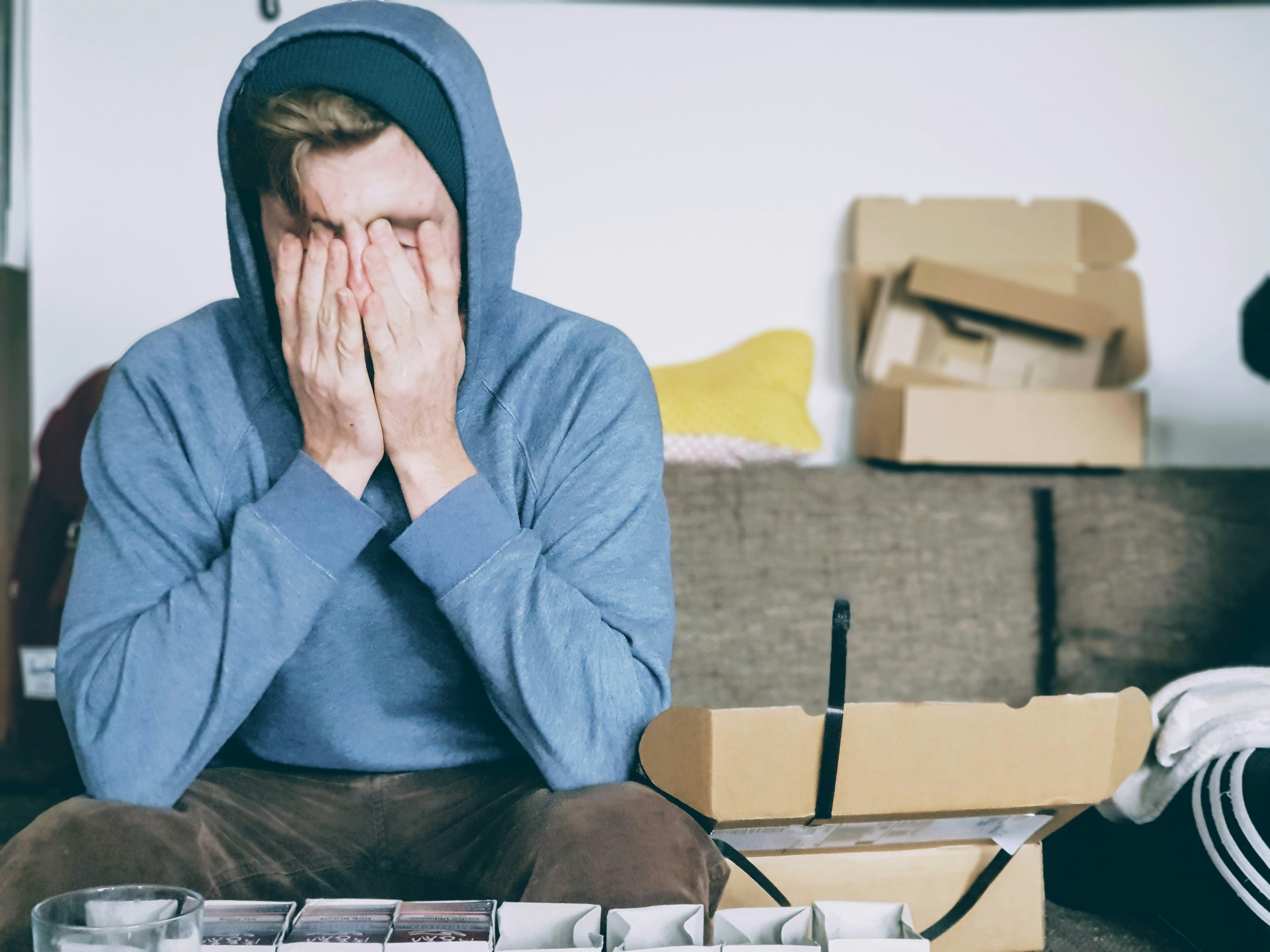 A picture of a man in a hoody sitting on a couch with his hands over his face, looking tired, in a messy room full of empty cardboard boxes.